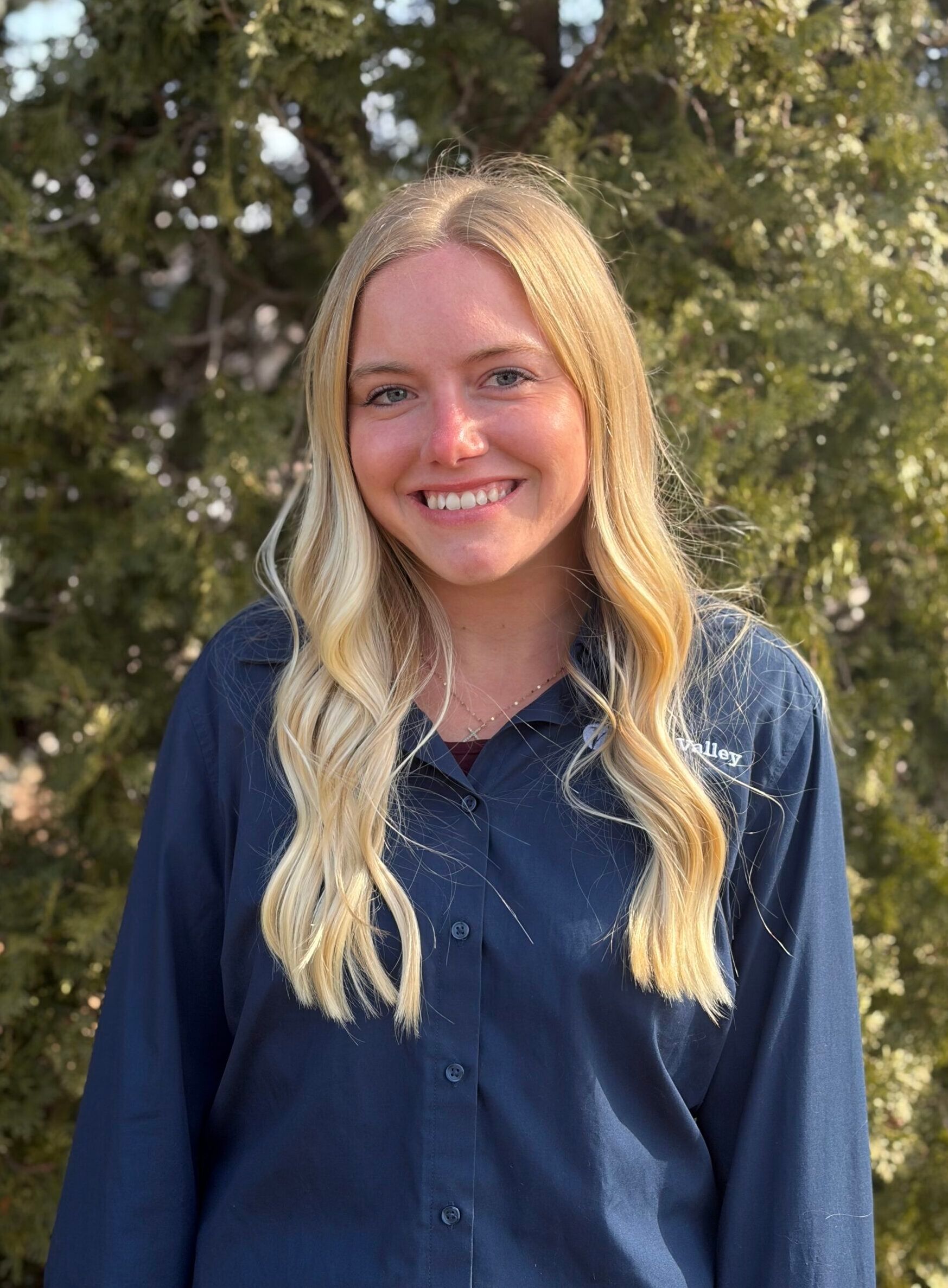 Smiling person in a navy shirt standing outdoors with trees in the background