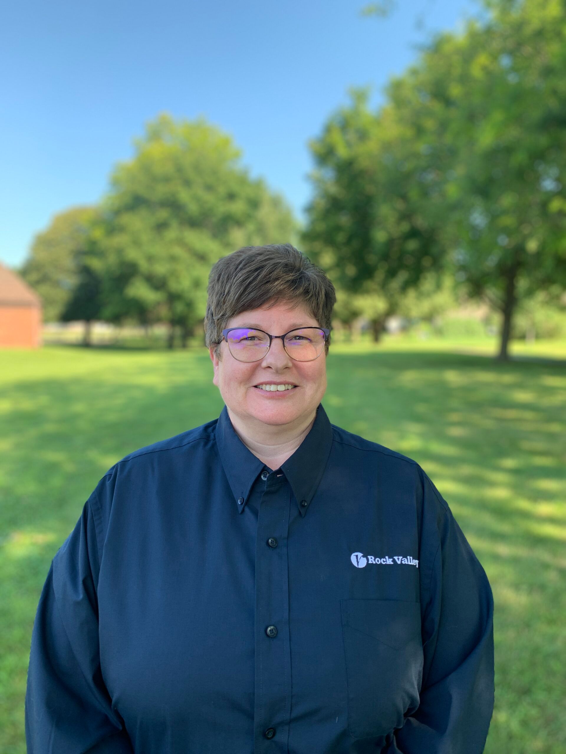 Smiling person in a navy shirt standing outdoors in a sunny park with green trees and blue sky.