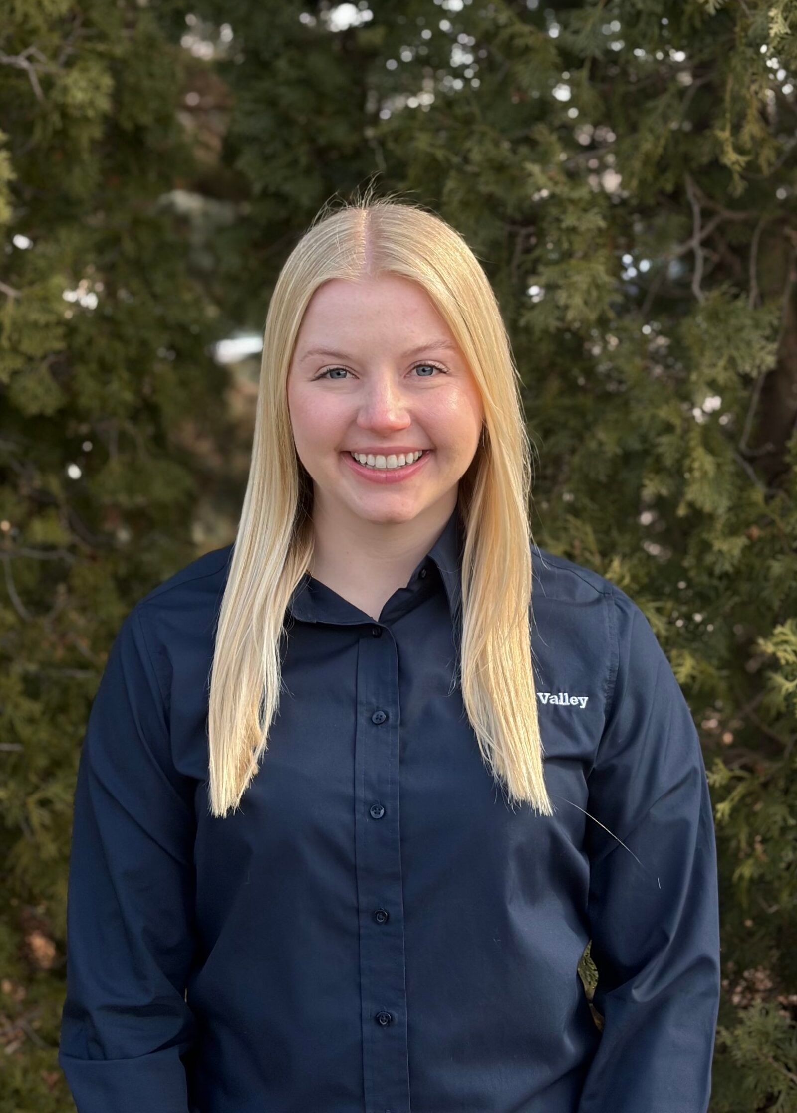 A person with long blonde hair and a friendly smile, wearing a dark blue button-down shirt outdoors in front of a tree.
