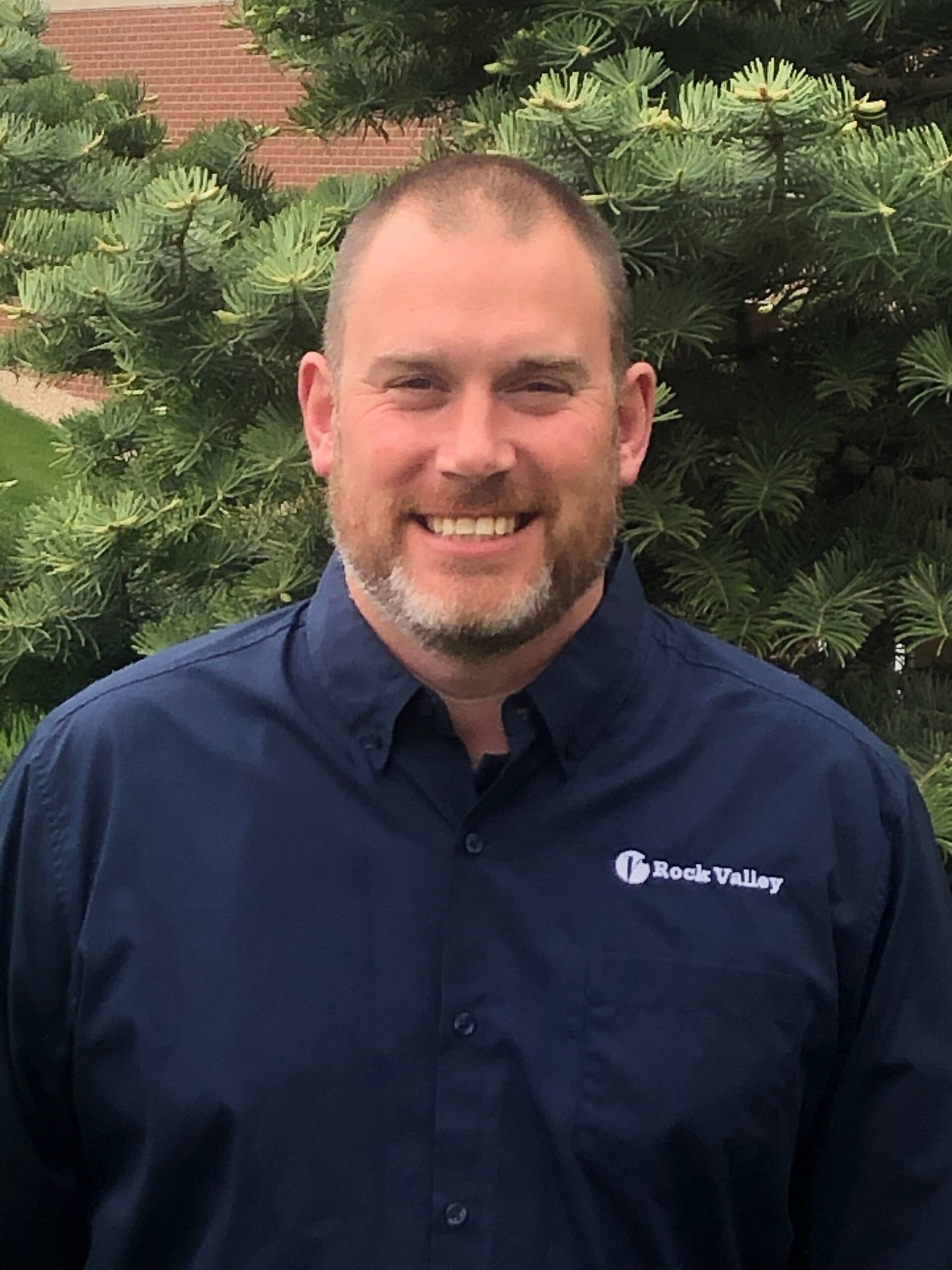 Smiling man in a navy shirt standing outdoors in front of green shrubs