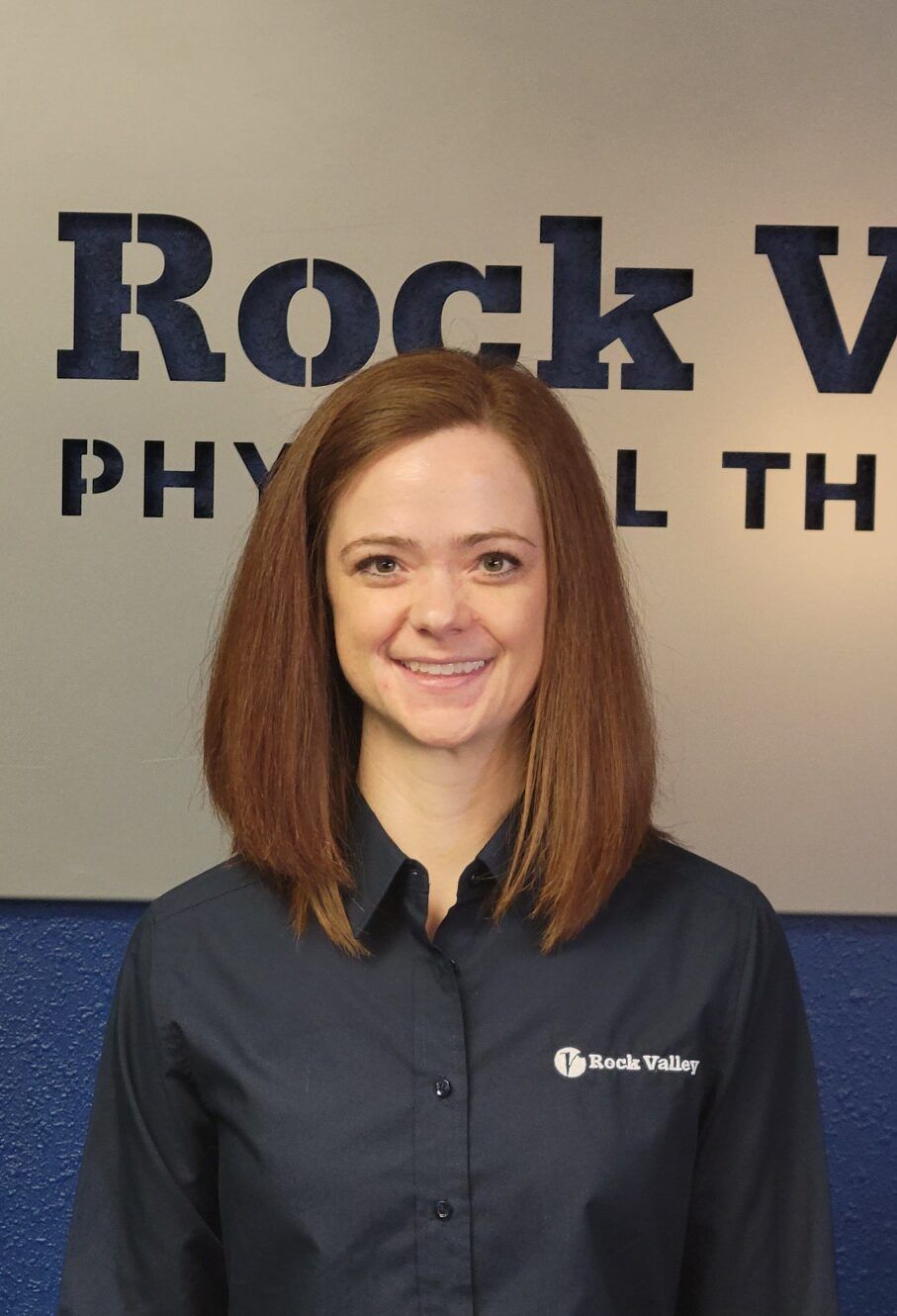 Woman smiling in front of a Rock Valley Physical Therapy sign, wearing a dark blue shirt.