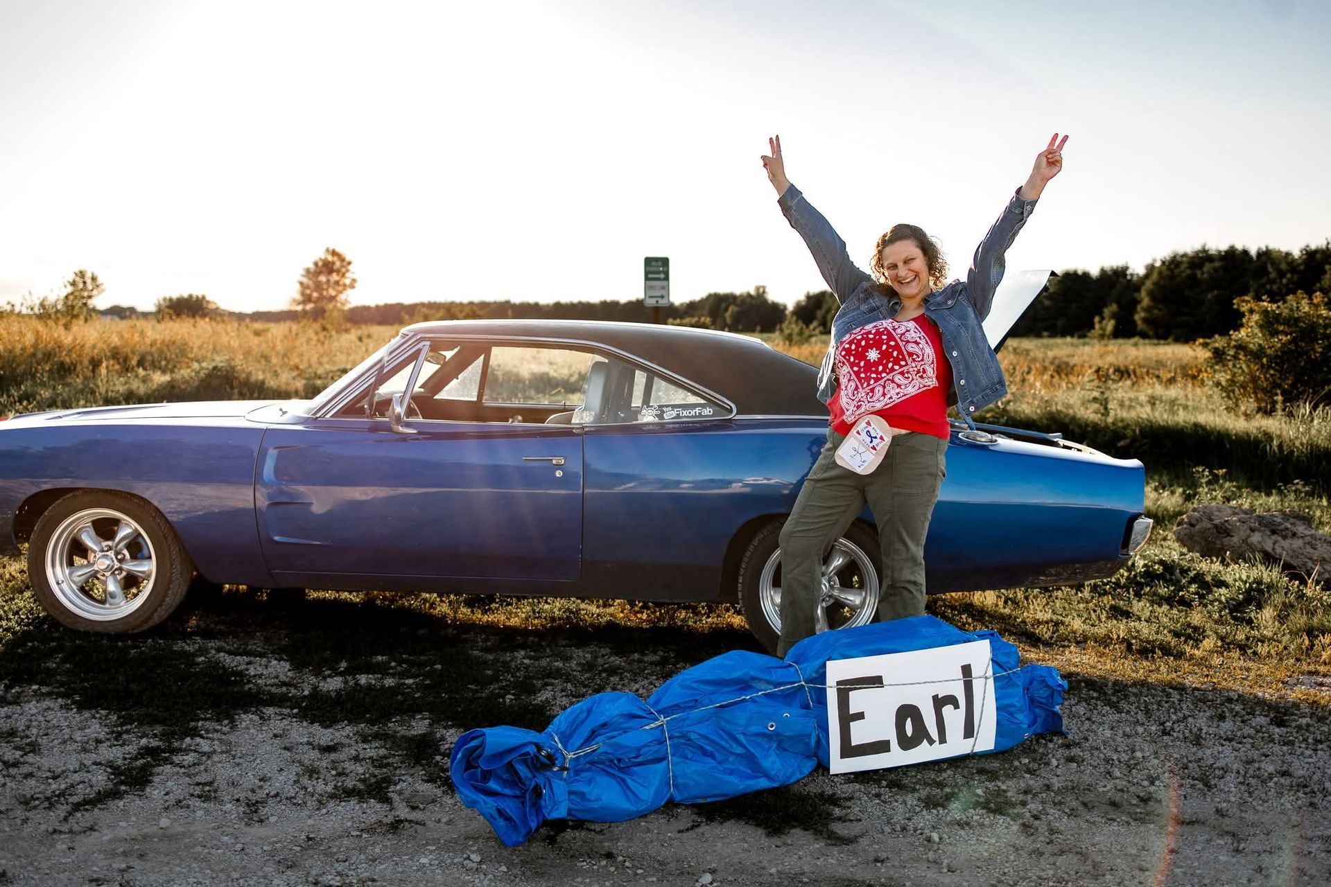Person posing beside a blue car with a sign reading “Earl” on the ground.