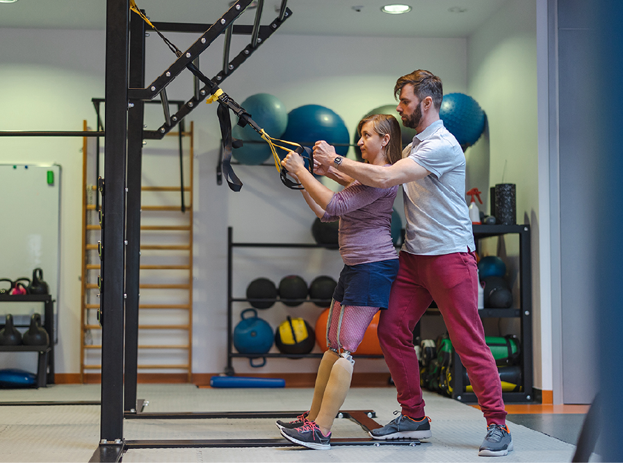 Woman with prosthetic legs working out with a physical therapist using suspension straps in a gym.