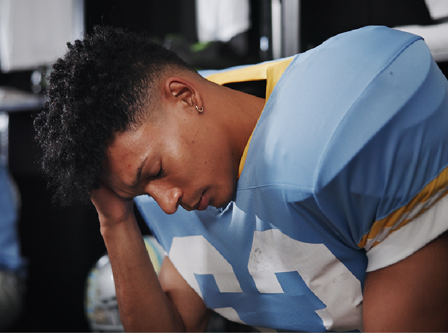 Football player in blue jersey sits hunched with head in hand, looking exhausted or disappointed