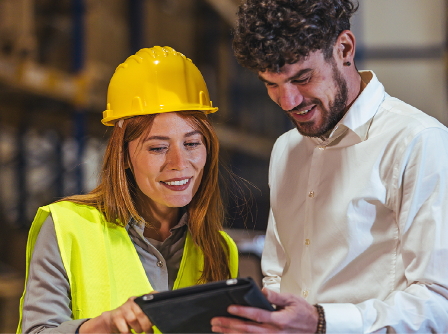 Two workers reviewing a tablet at a construction site, one wearing a yellow hard hat and safety vest.