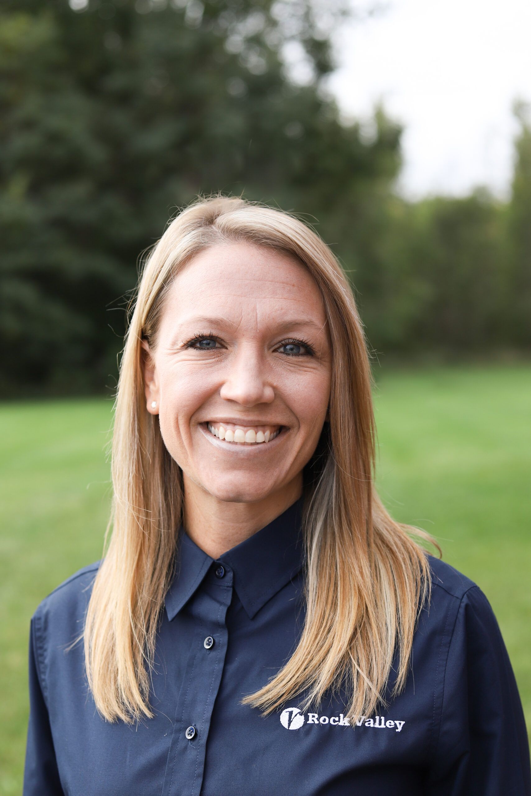 Woman with blonde hair smiles at the camera, wearing a blue button-down shirt outdoors.