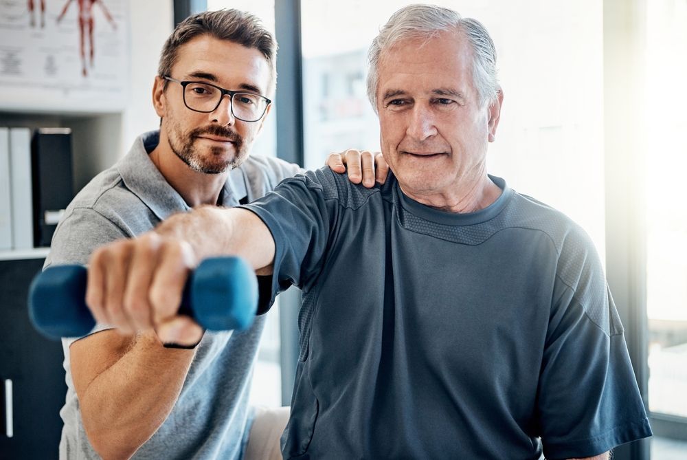 Two men in a bright room, one holding a blue dumbbell in front, suggesting physical therapy or exercise