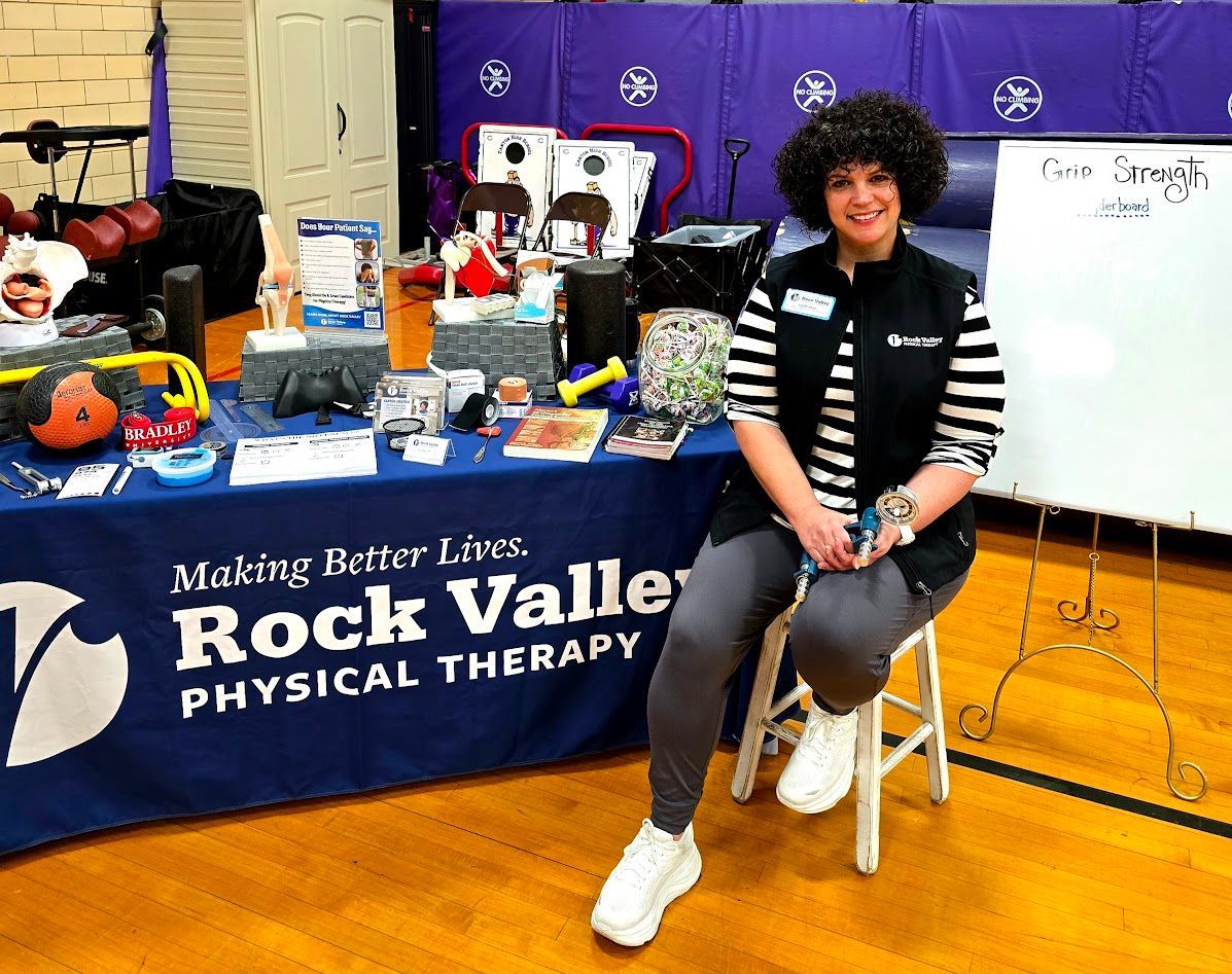 Woman seated at a Rock Valley Physical Therapy booth, with display table and whiteboard in a gym.