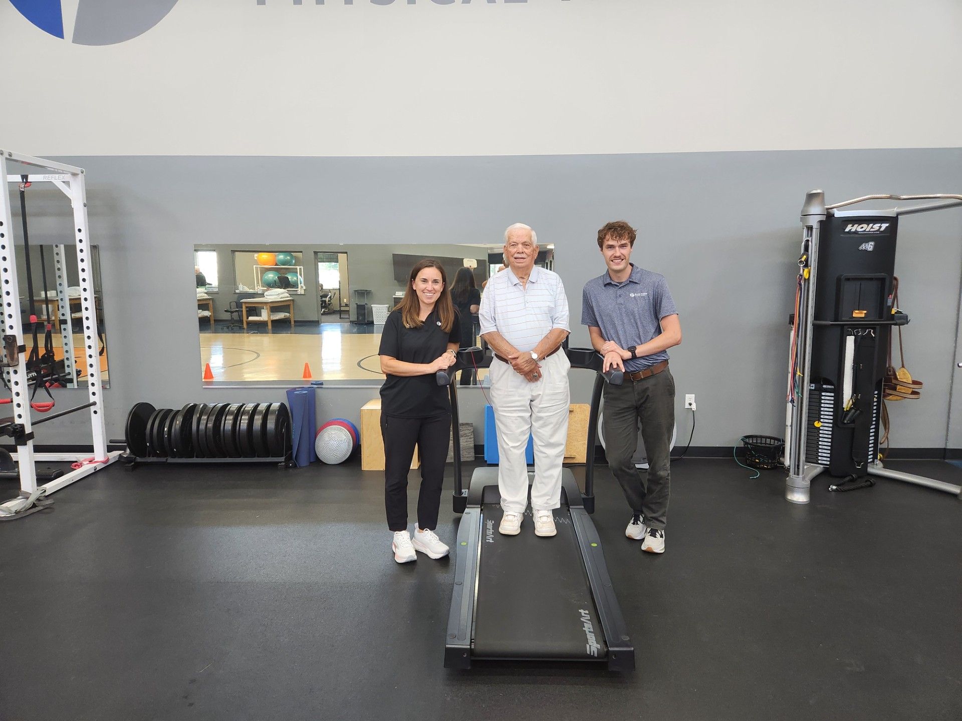 Three people standing on a treadmill in a gym, with exercise equipment in the background.
