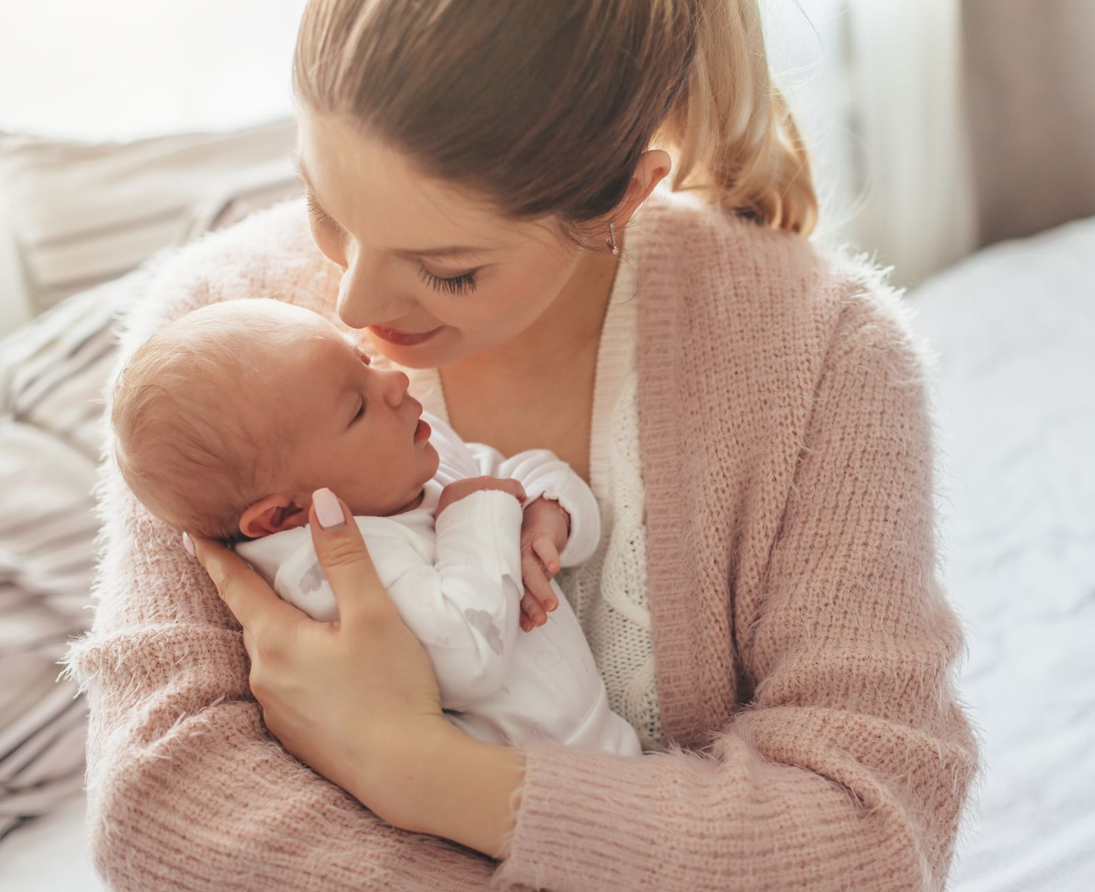 Woman holding a newborn baby on a bed, leaning in affectionately near a sunlit window.