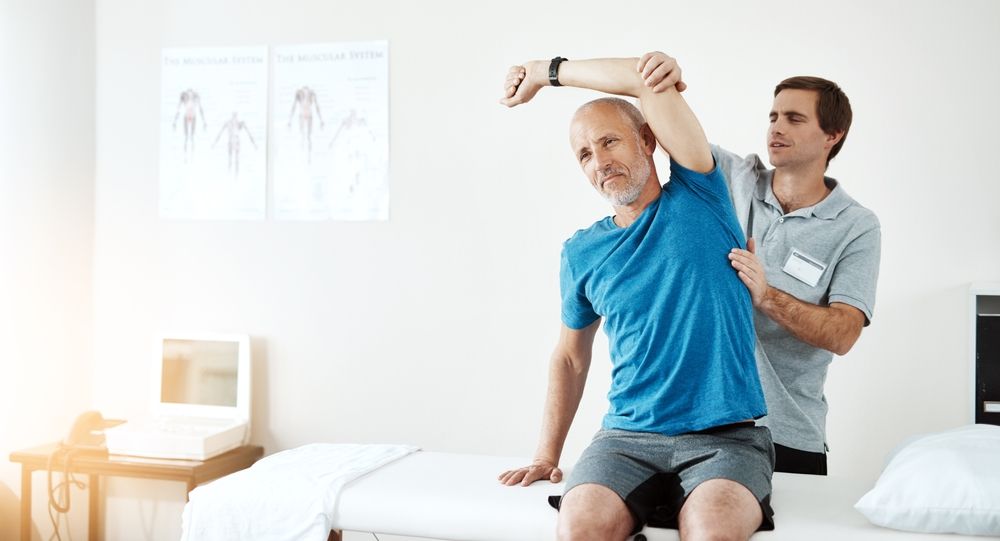 Physical therapist helping a seated patient stretch his arm in a bright clinic room