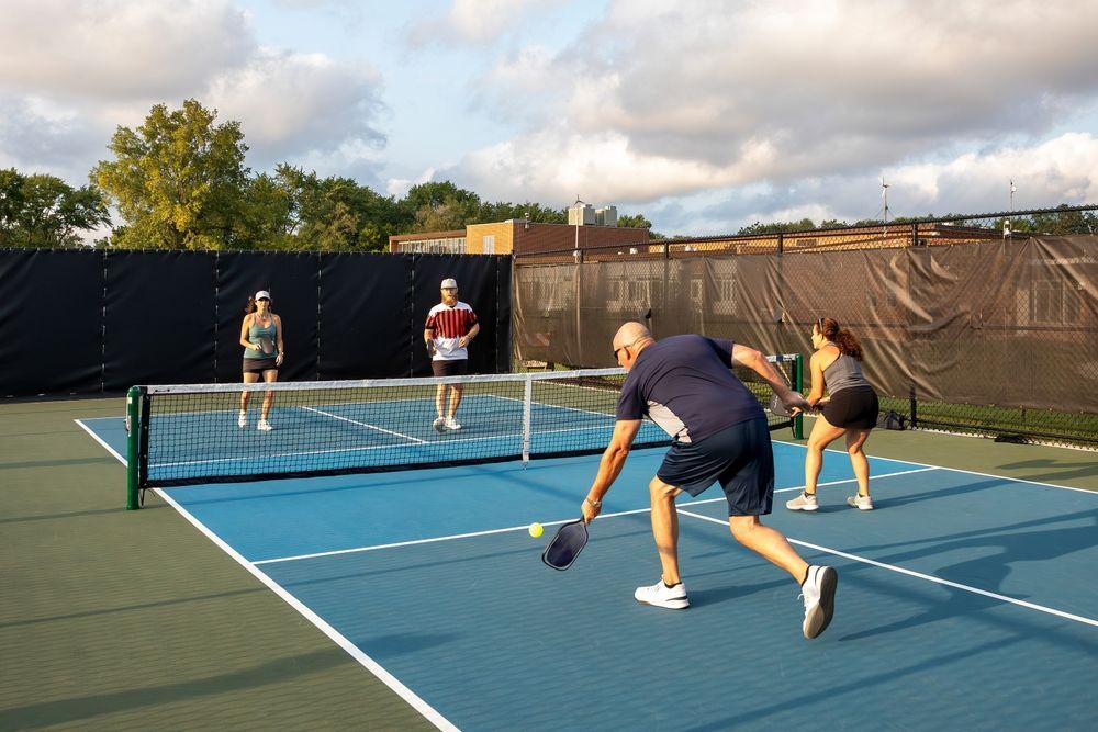 People playing pickleball on a blue outdoor court, with a black net and fence in the background