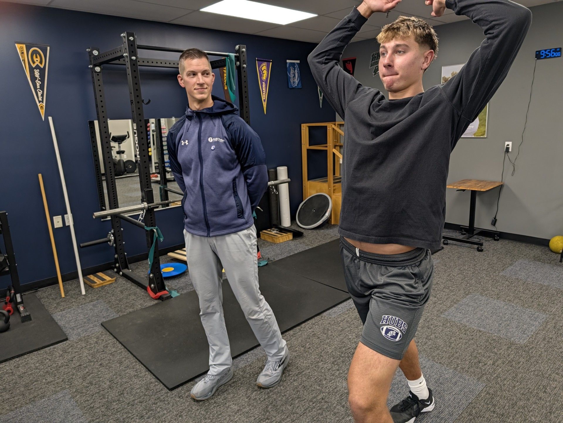 Two men exercising in a gym, one stretching with hands overhead and one standing beside him.