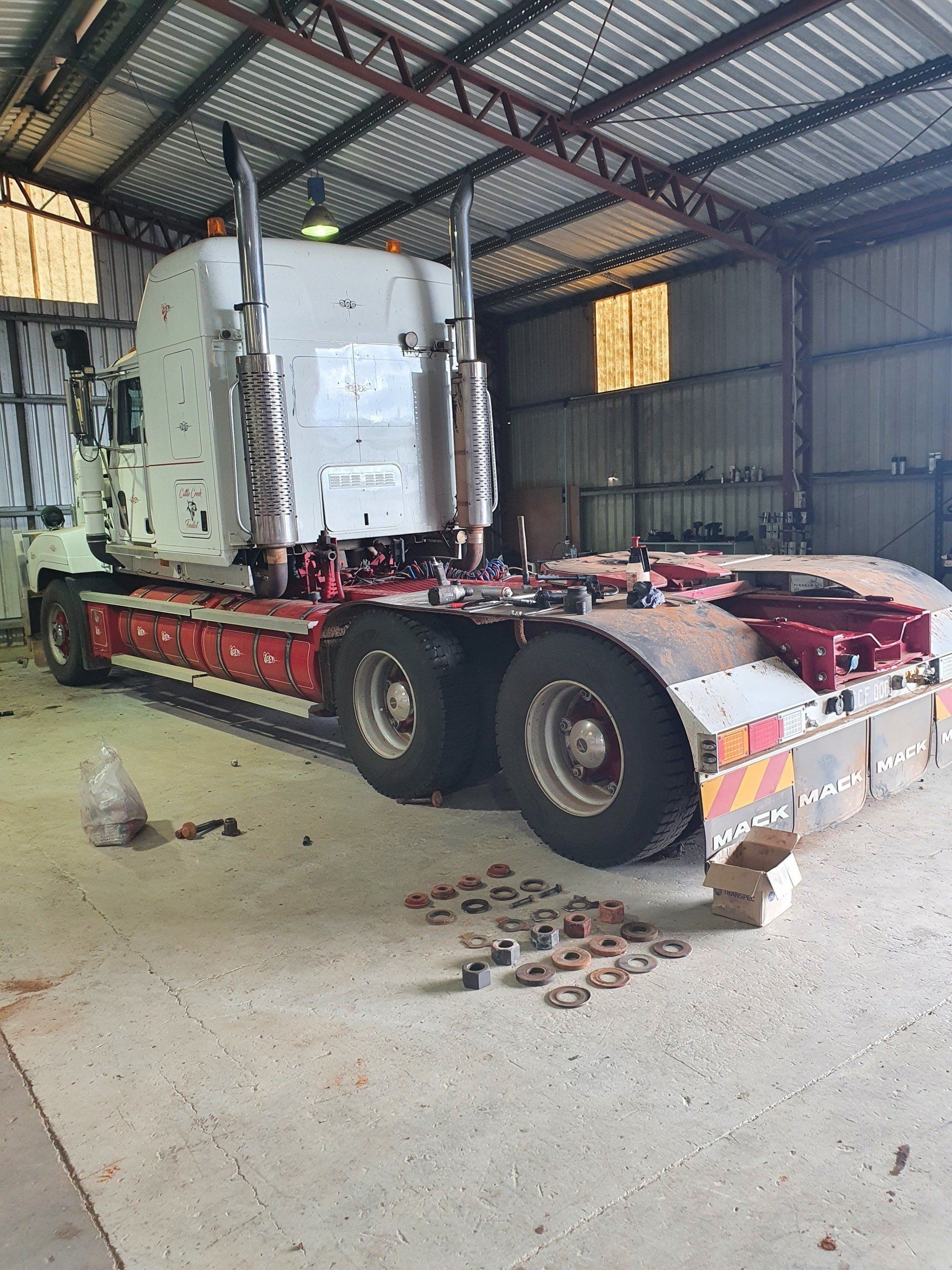 White semi-truck inside a garage with scattered parts on the floor; red accents, metal structure.