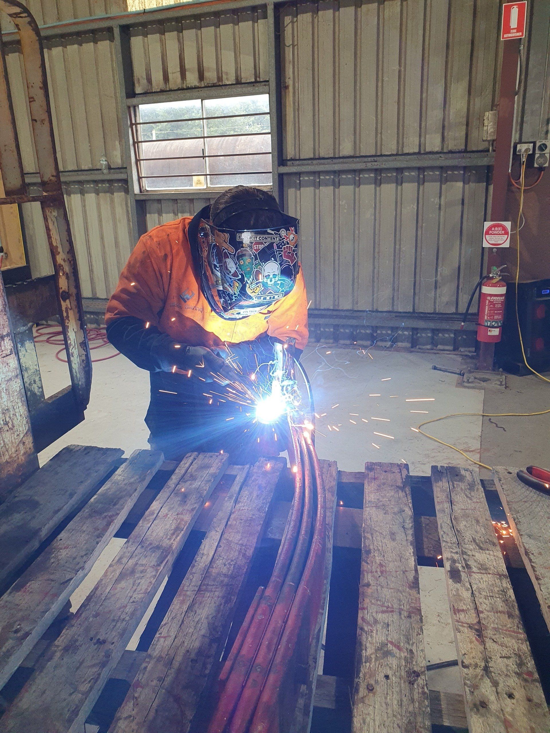 Welder in protective gear welding metal pieces on a wooden pallet inside a workshop.