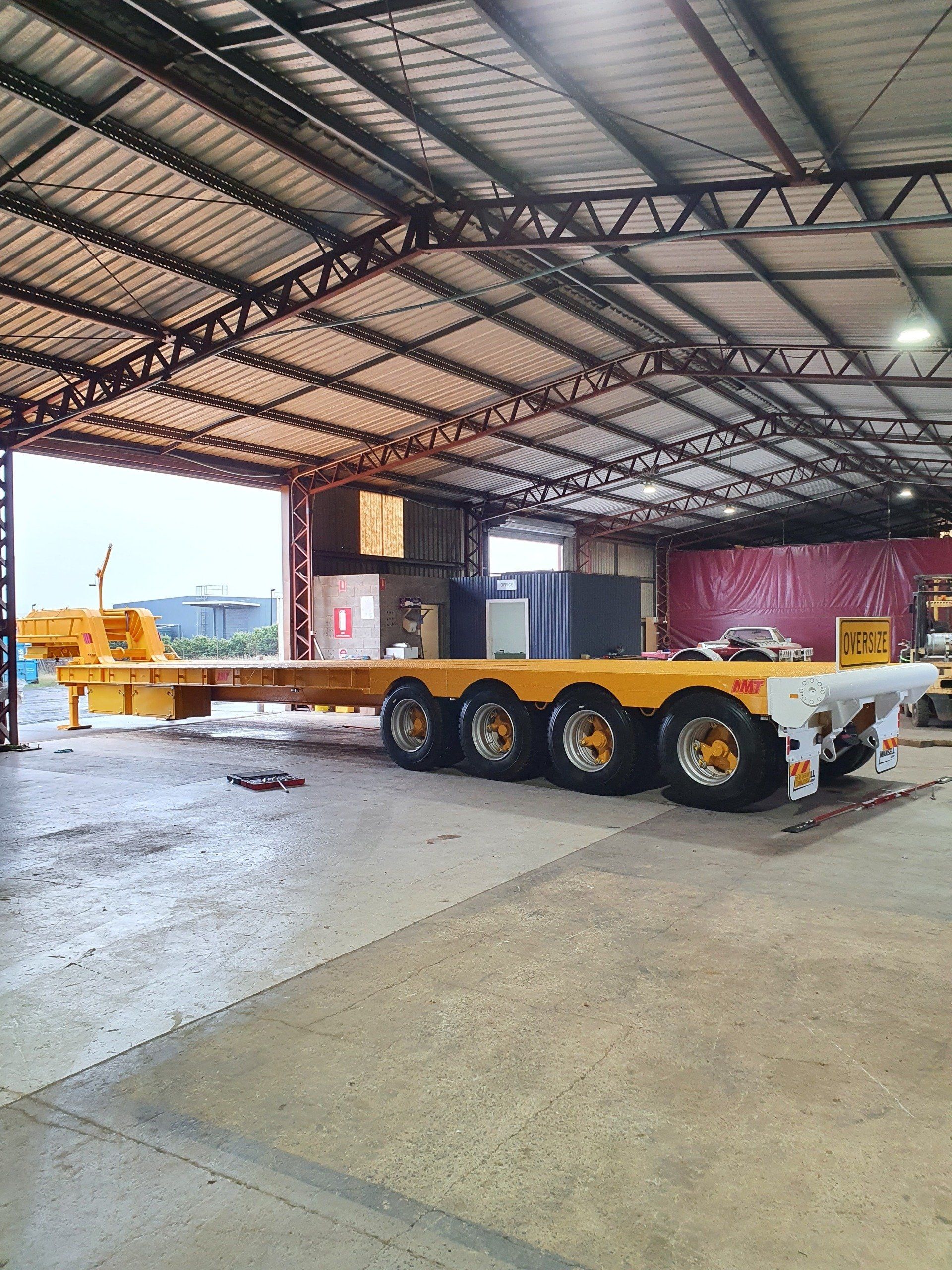 Yellow flatbed trailer inside a warehouse, with several axles and wheels.