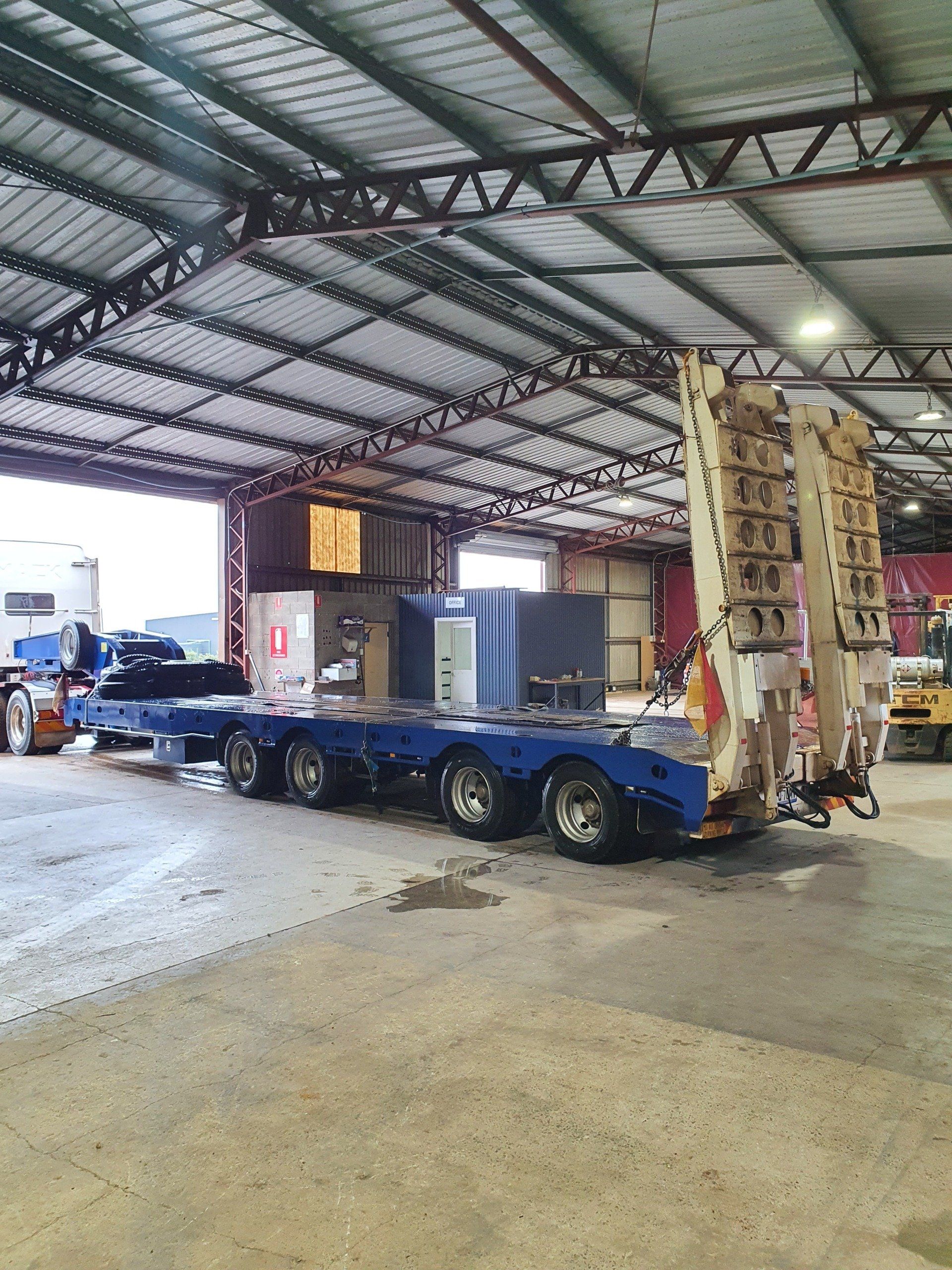 Large, blue semi-truck trailer carrying equipment inside a warehouse. Steel beams overhead.