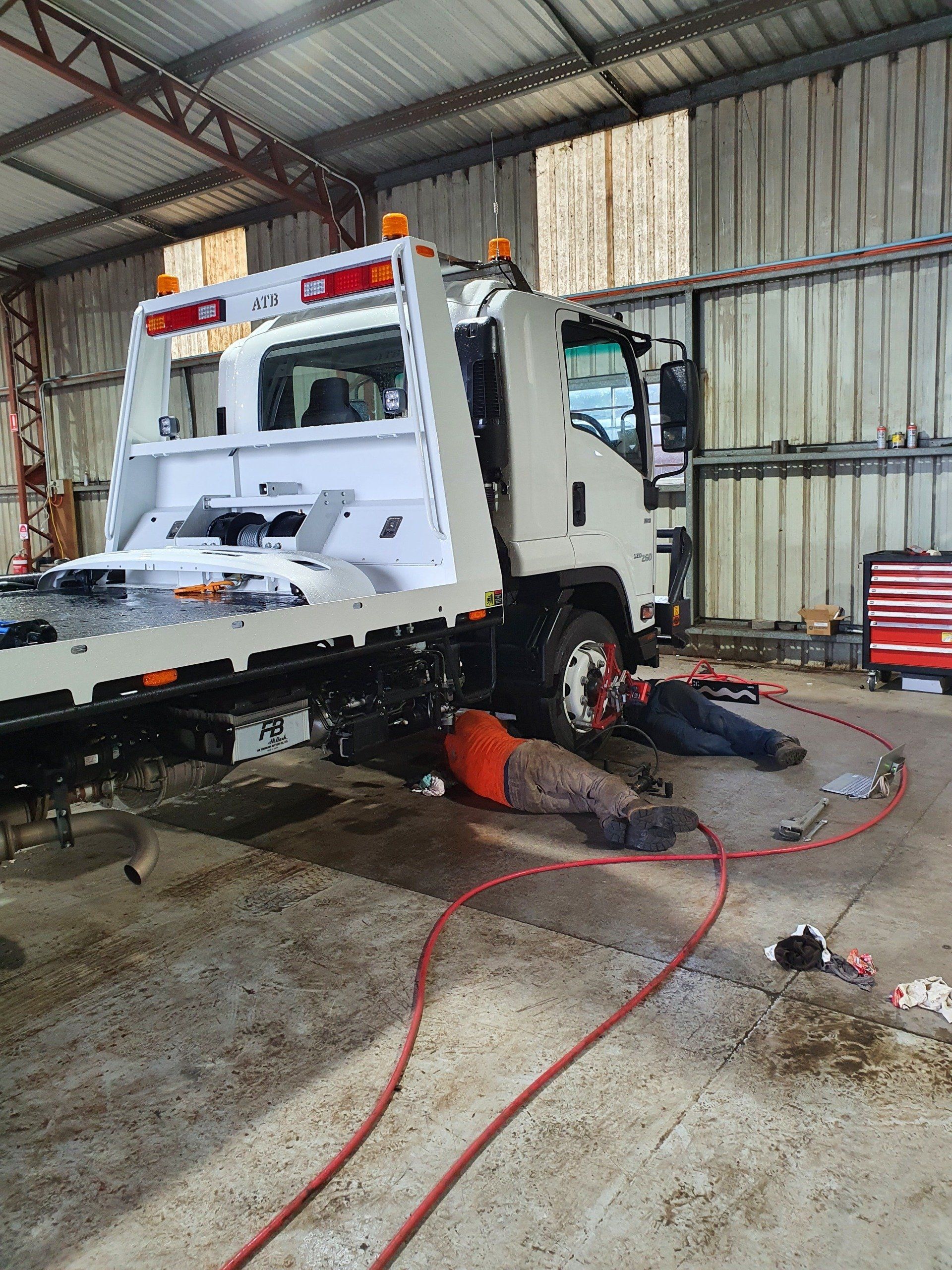 Tow truck in a shop, mechanic working on the front wheel, red hoses, tools visible, interior setting.