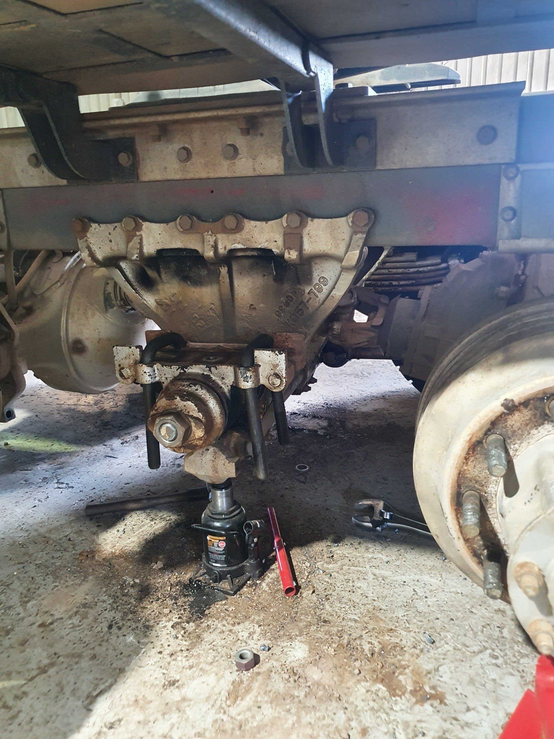 Undercarriage of a truck being worked on. A jack supports the axle. Tools and parts are scattered around the area.