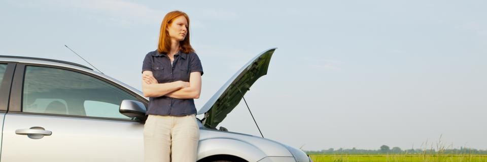 Person standing with arms crossed beside a silver car with its hood open in a field.