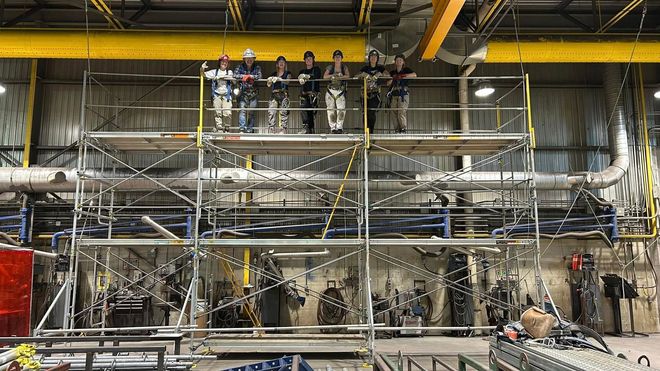 Des ouvriers travaillent sur des échafaudages à l'intérieur d'une grande installation industrielle, avec des tuyaux et des équipements jaunes suspendus au plafond.