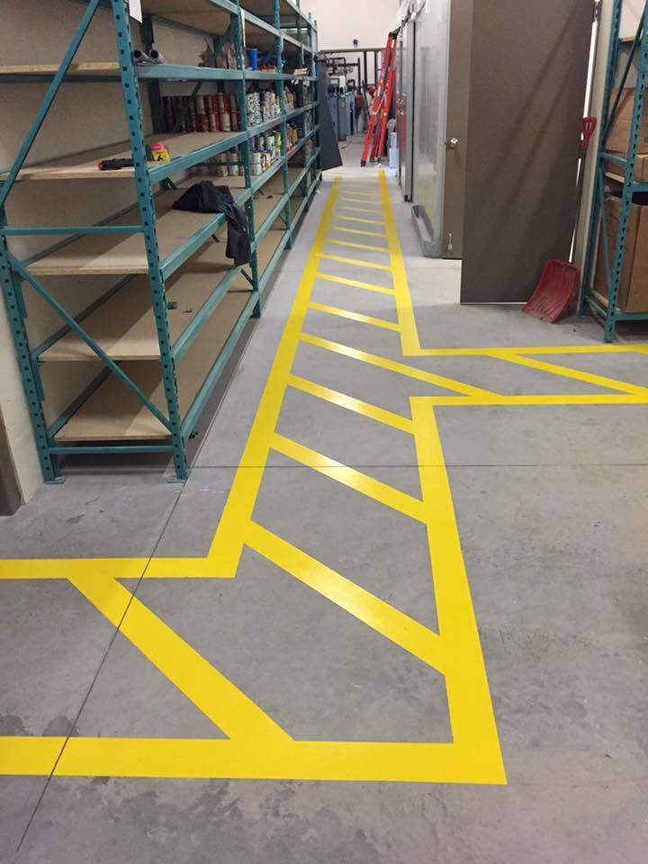Allée d'entrepôt avec marquages au sol jaune vif et étagères métalliques sur le côté gauche.