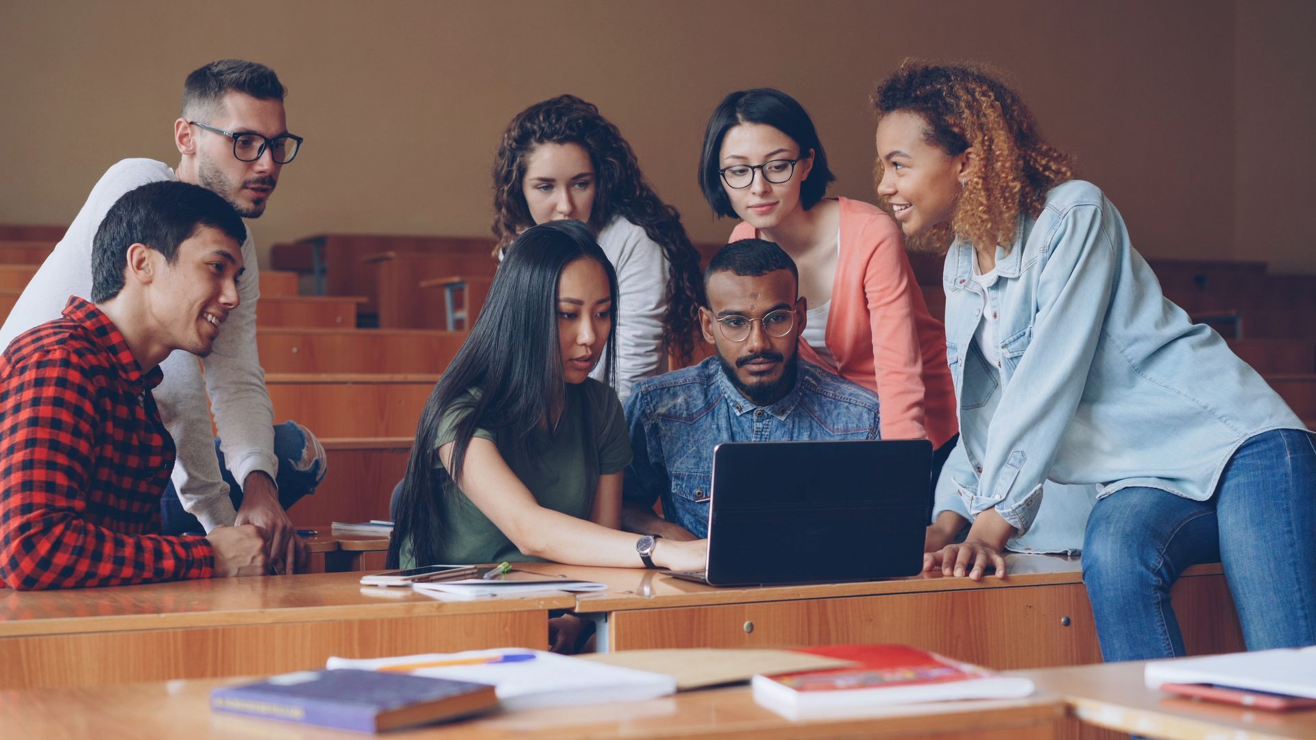 A diverse group of students in a lecture hall looking at a laptop screen together. A diverse group of students in a lecture hall looking at a laptop screen together.