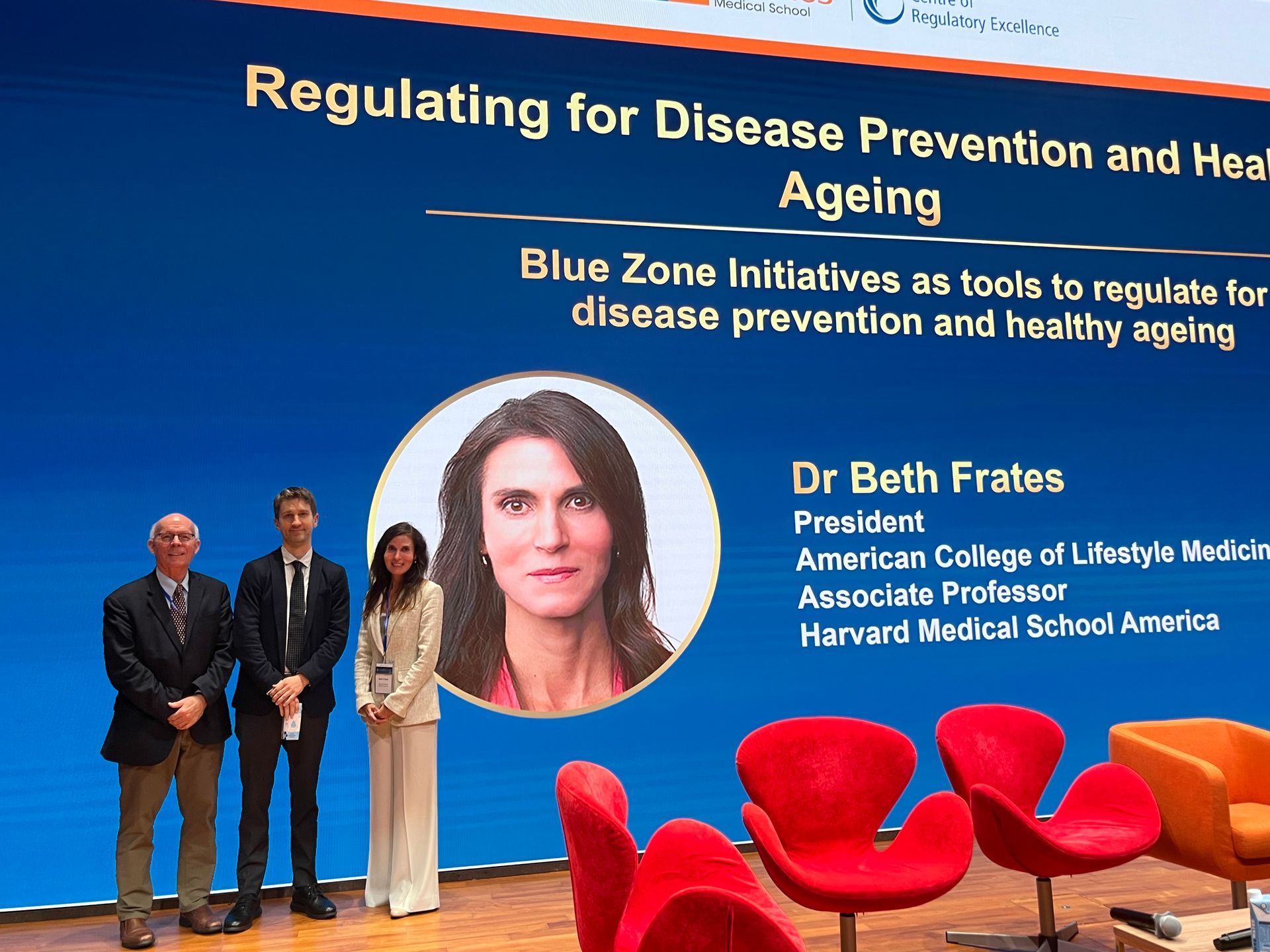 Conference slide on disease prevention and healthy ageing with speaker photo and panel on stage