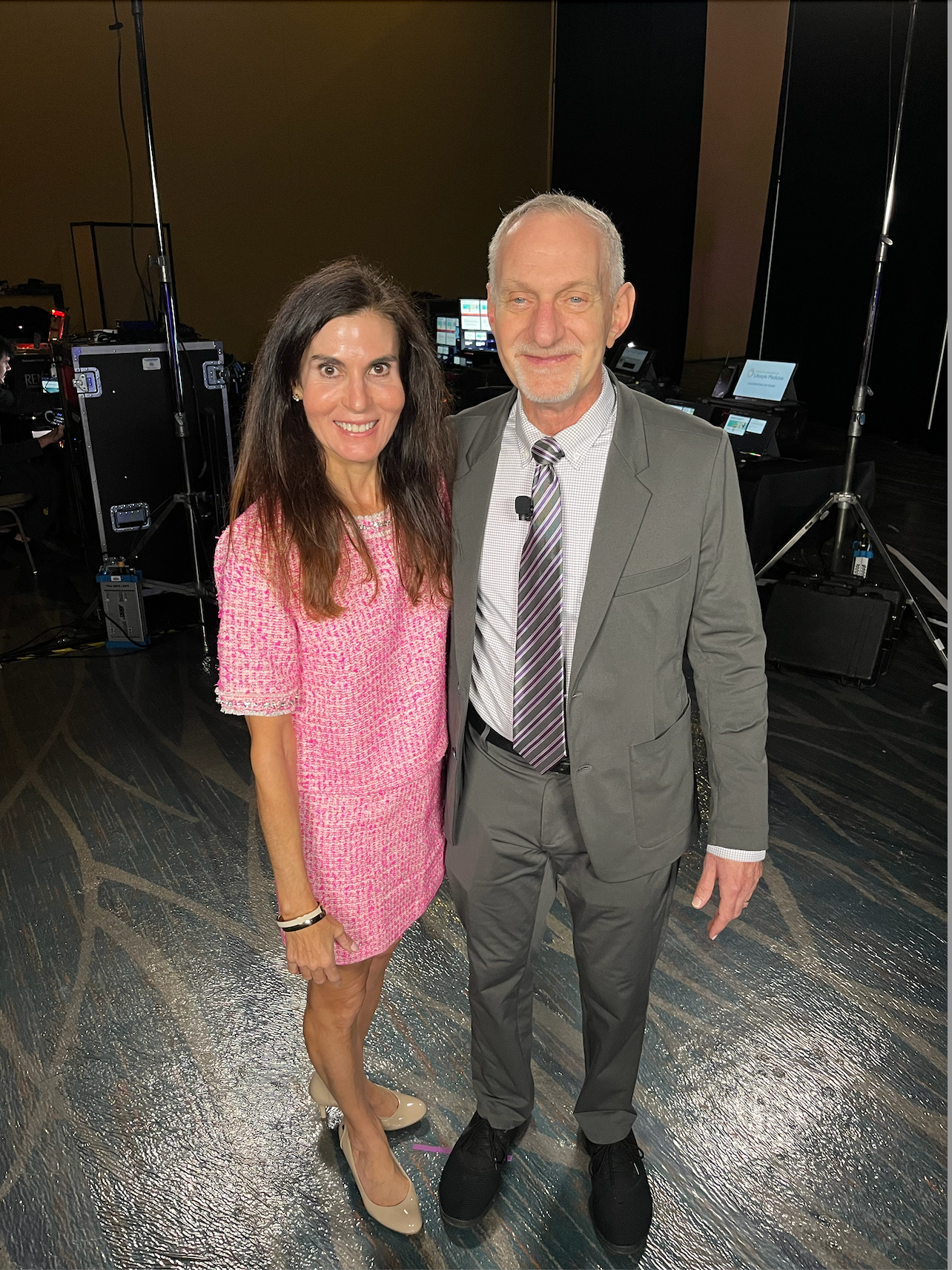 Two people posing on a stage, one in a pink dress and one in a gray suit, with studio lights behind them
