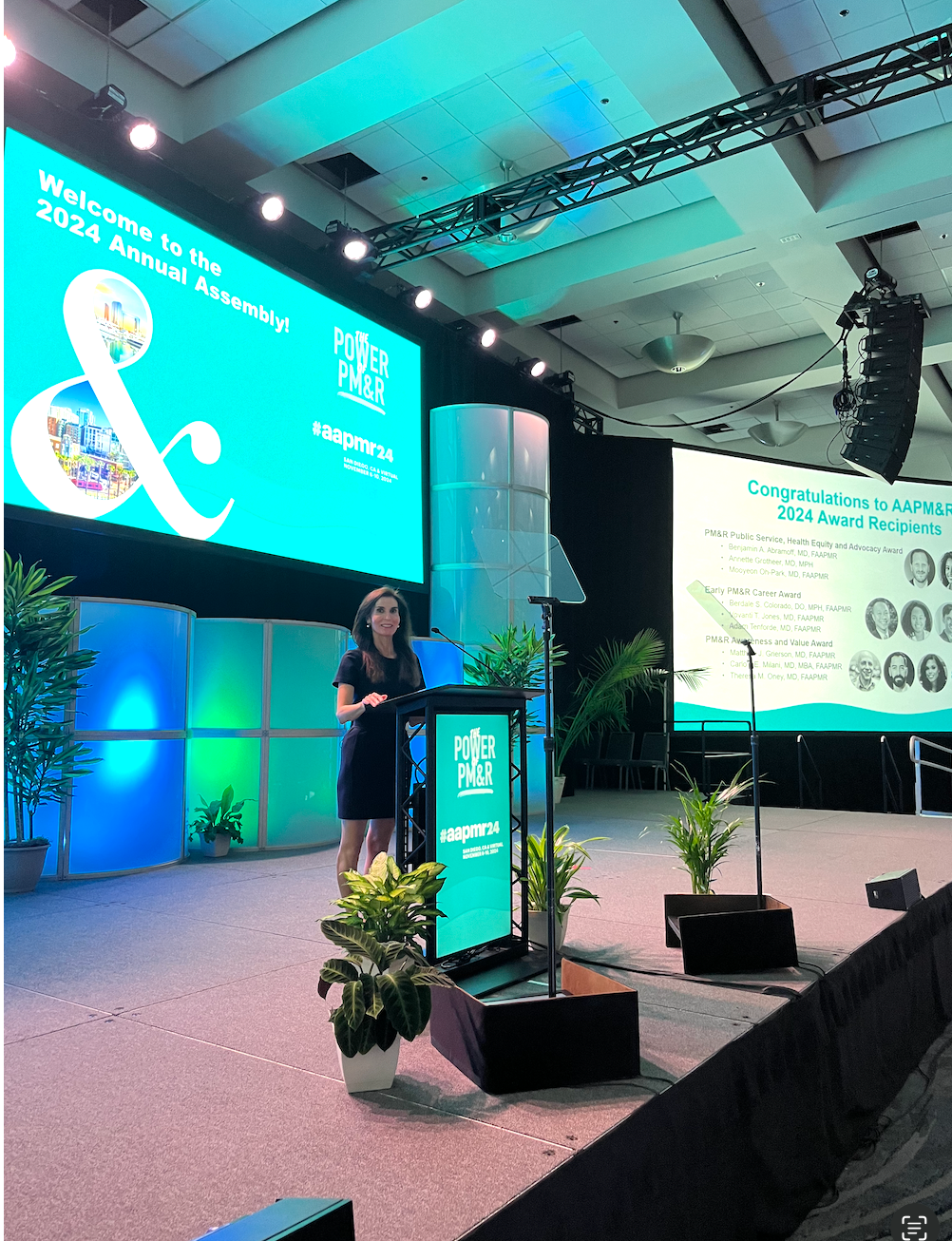 Speaker at podium on a lit conference stage with large presentation screens and potted plants