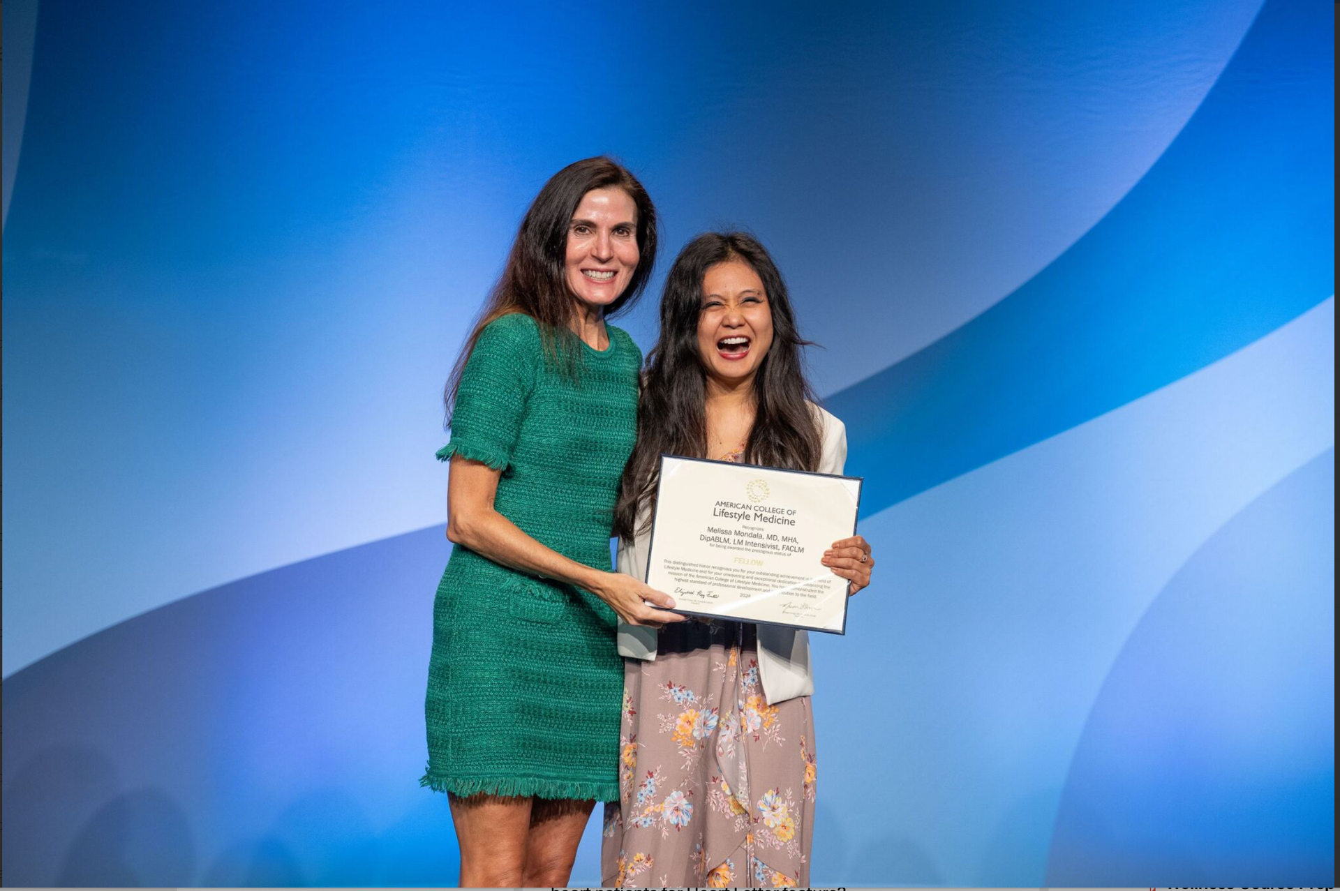 Two people smiling onstage, holding a certificate against a blue backdrop.