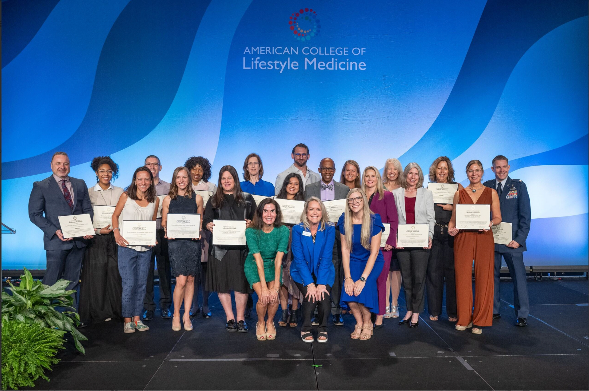 Group of people onstage holding certificates at an awards event with a blue backdrop