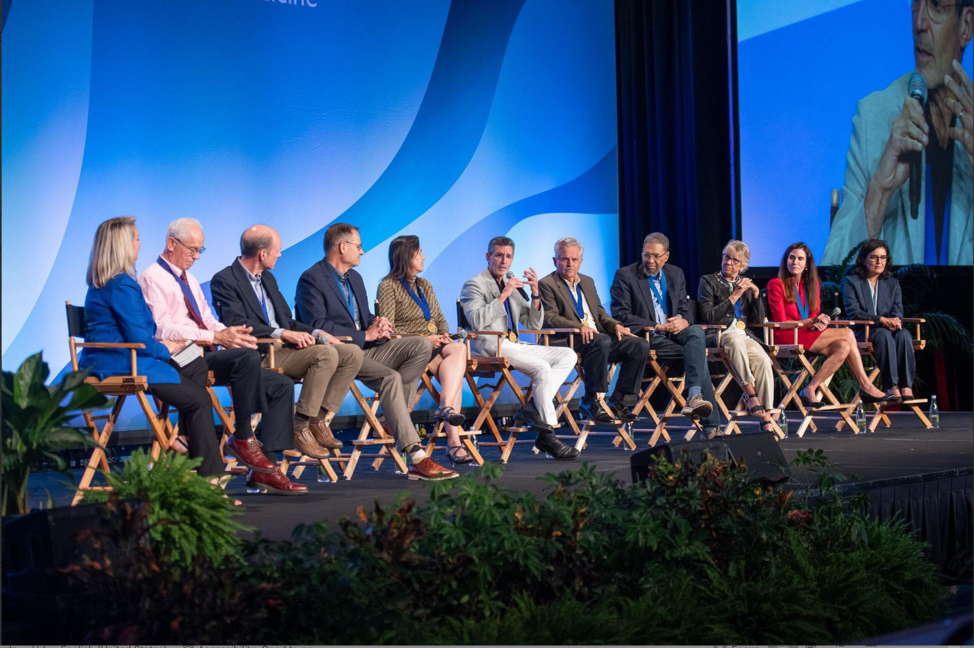 Panel of speakers seated onstage at a conference, with blue lighting and a large projected image behind them.