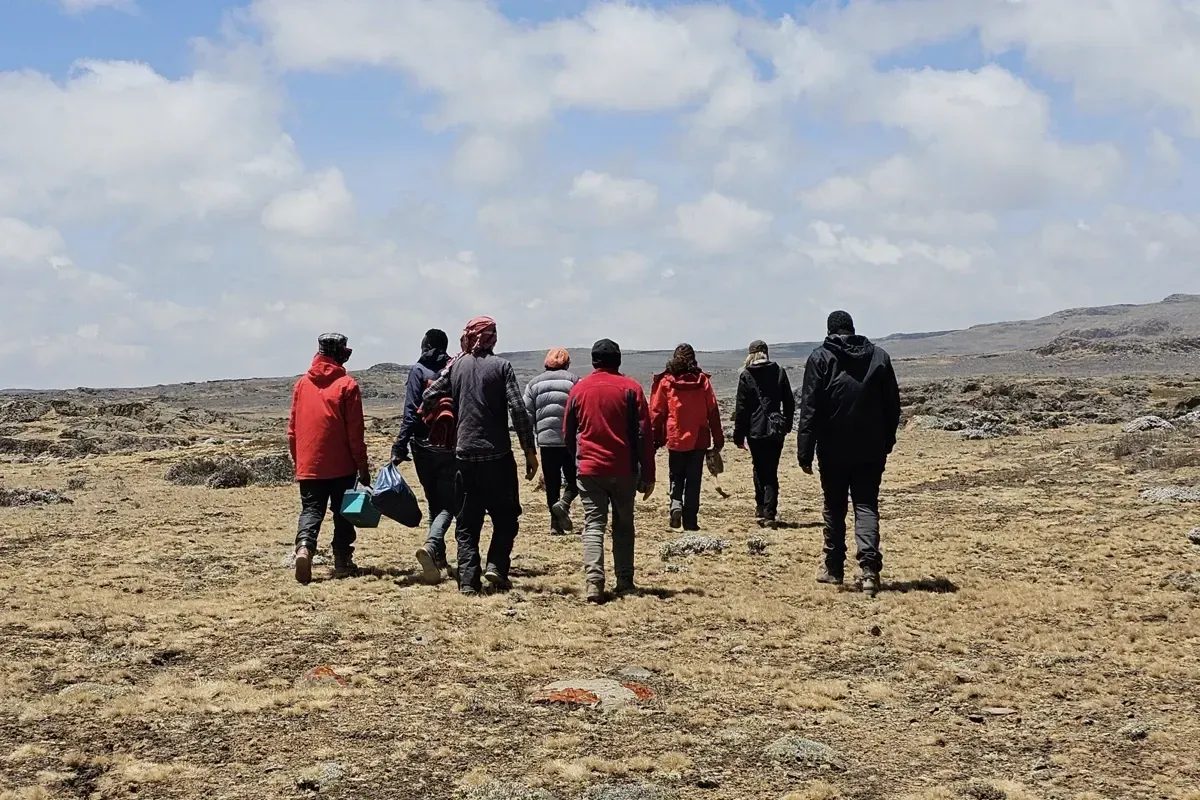 Group of hikers in red and dark jackets walking across a rocky, barren landscape under a cloudy sky