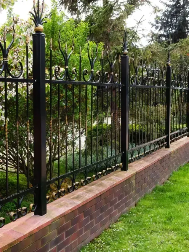 El monte fence company - Black wrought iron fence topped with intricate designs, set atop a brick wall, surrounded by lush greenery.