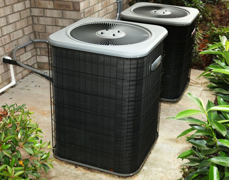 Two black air conditioning units outdoors near greenery and a brick wall.