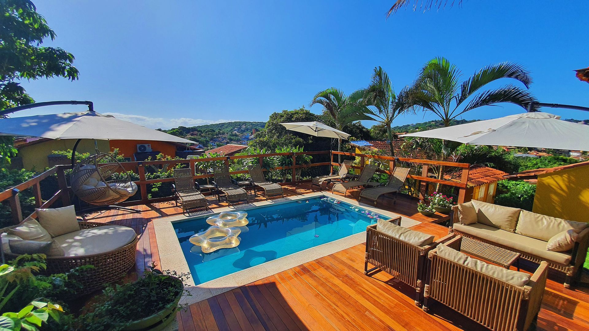 Pátio tropical nos fundos da casa com piscina azul, deck de madeira, espreguiçadeiras, guarda-sóis e palmeiras.