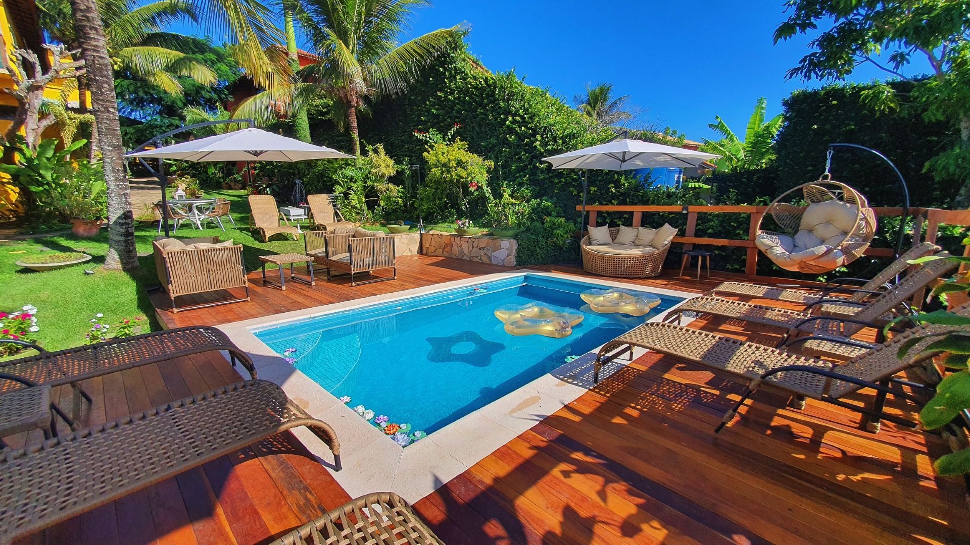 Terraço tropical à beira da piscina com espreguiçadeiras, guarda-sóis e vegetação exuberante sob um céu azul brilhante.
