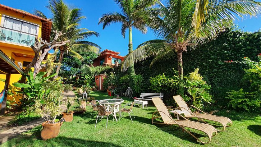 Jardim tropical ensolarado com palmeiras, espreguiçadeiras e vegetação exuberante ao lado de uma casa colorida.