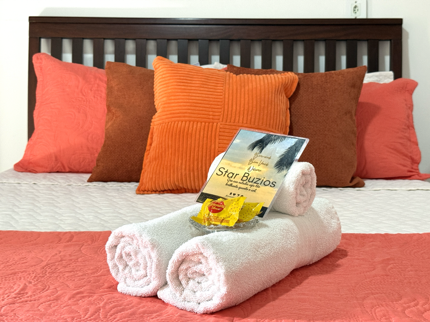 Toalhas brancas dobradas sobre uma cama com travesseiros corais e laranjas, com uma pequena sacola de presente e artigos de higiene pessoal por cima.