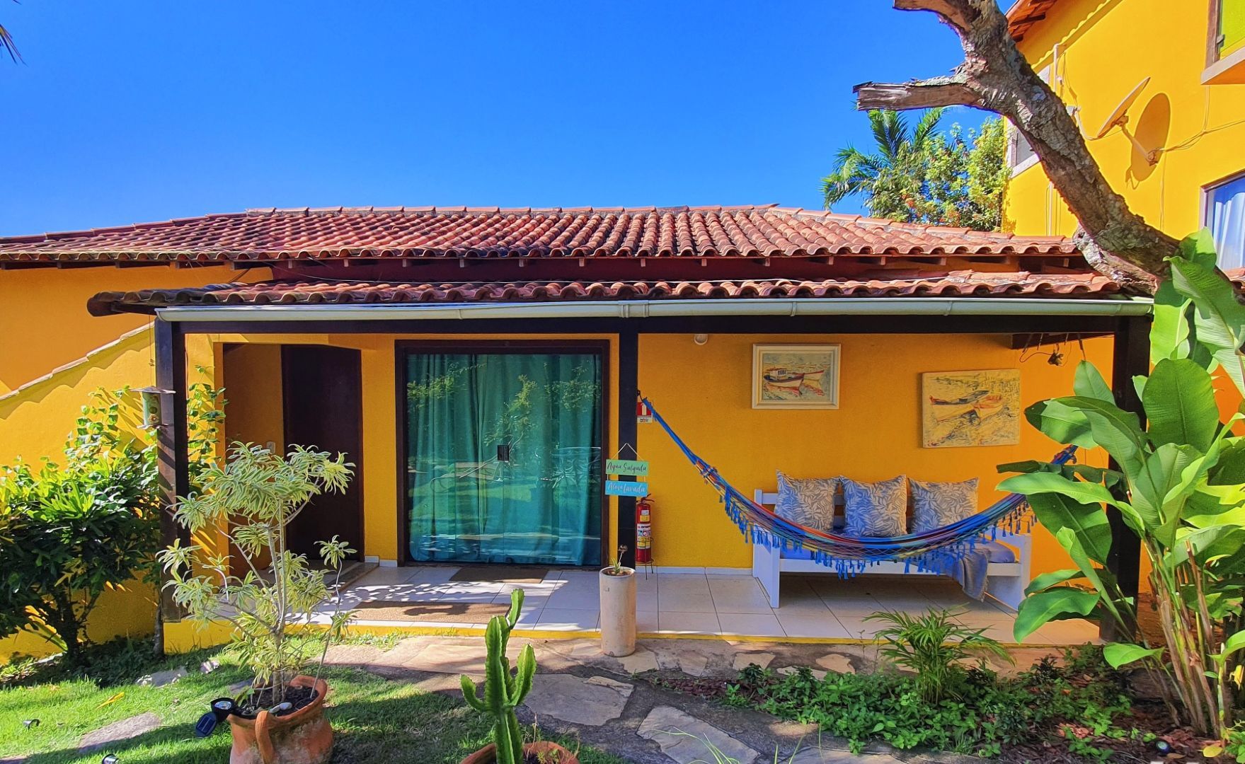 Pátio de casa amarela com cortinas azuis, telhado de telhas e plantas tropicais exuberantes em primeiro plano.