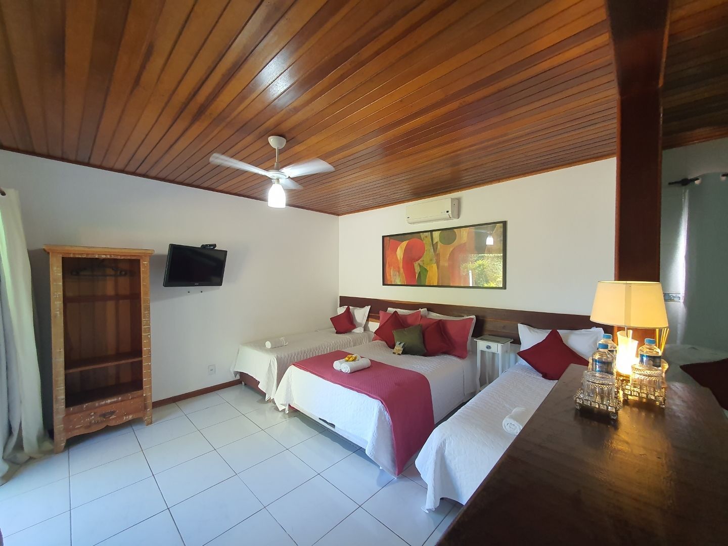 Quarto com duas camas de solteiro, teto de madeira, TV de parede e iluminação aconchegante com abajur ao lado da cama.