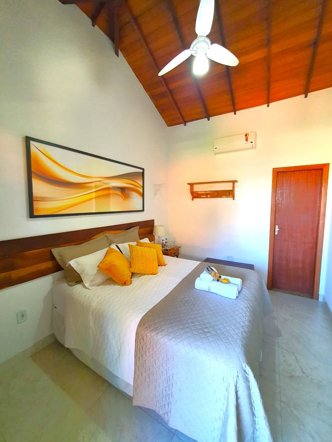 Quarto pequeno com cama, lençóis brancos, almofadas amarelas, decoração de parede, ventilador de teto e porta de madeira.