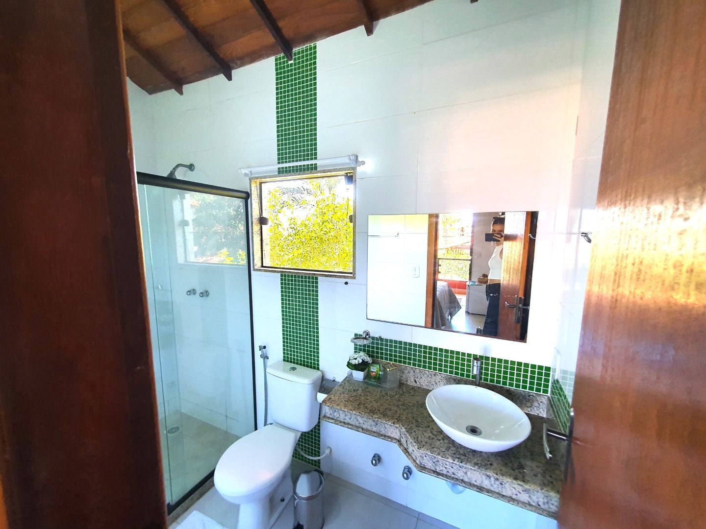 Banheiro compacto com vaso sanitário, box de vidro, pia e espelho em paredes revestidas com azulejos, sob um teto inclinado de madeira.