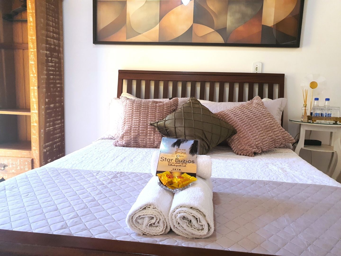 Cama arrumada com colcha branca, almofadas estampadas, toalhas enroladas e uma bandeja com bebidas de laranja em um quarto iluminado.