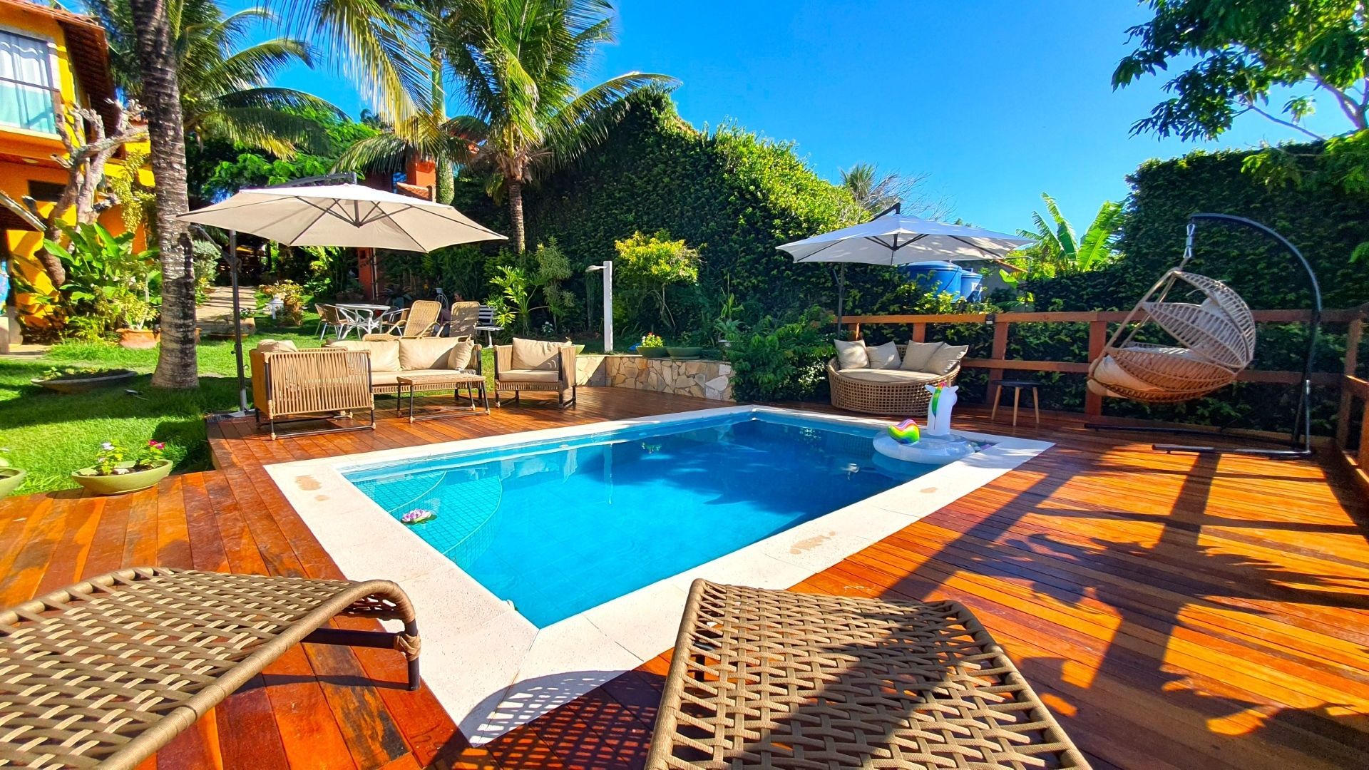 Pátio ensolarado nos fundos com uma pequena piscina azul, deck de madeira, espreguiçadeiras e guarda-sóis.