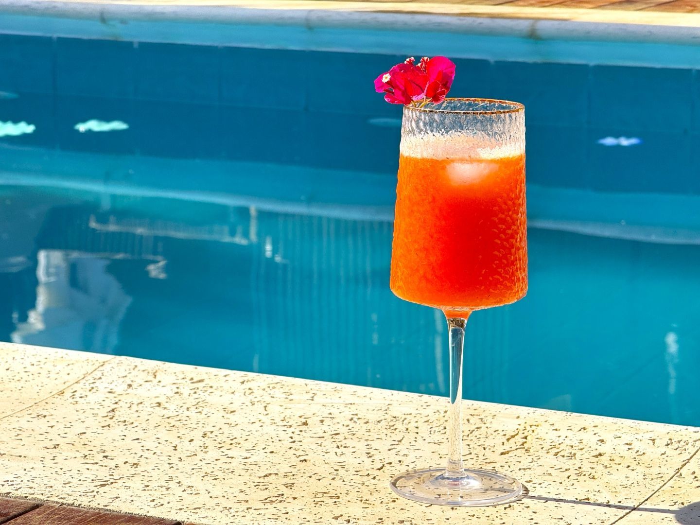 Coquetel de laranja servido em uma taça com haste, ao lado de uma piscina, decorado com uma flor rosa.
