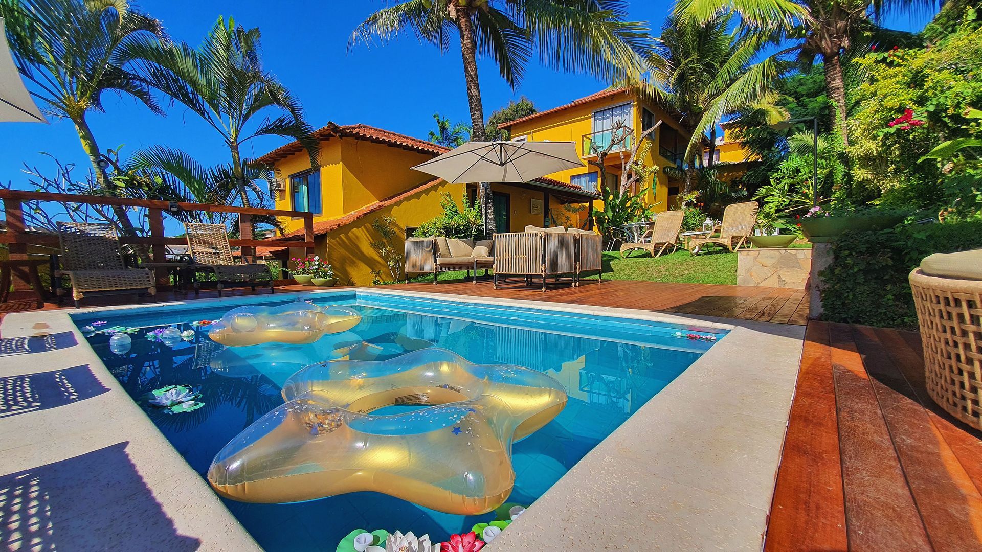 Piscina com cisne flutuante, casa amarela e palmeiras sob um sol tropical intenso.