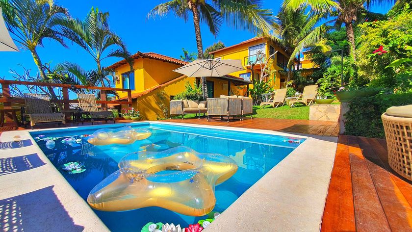 Piscina tropical iluminada com água azul, espreguiçadeiras, palmeiras e chalés amarelos sob um céu ensolarado.
