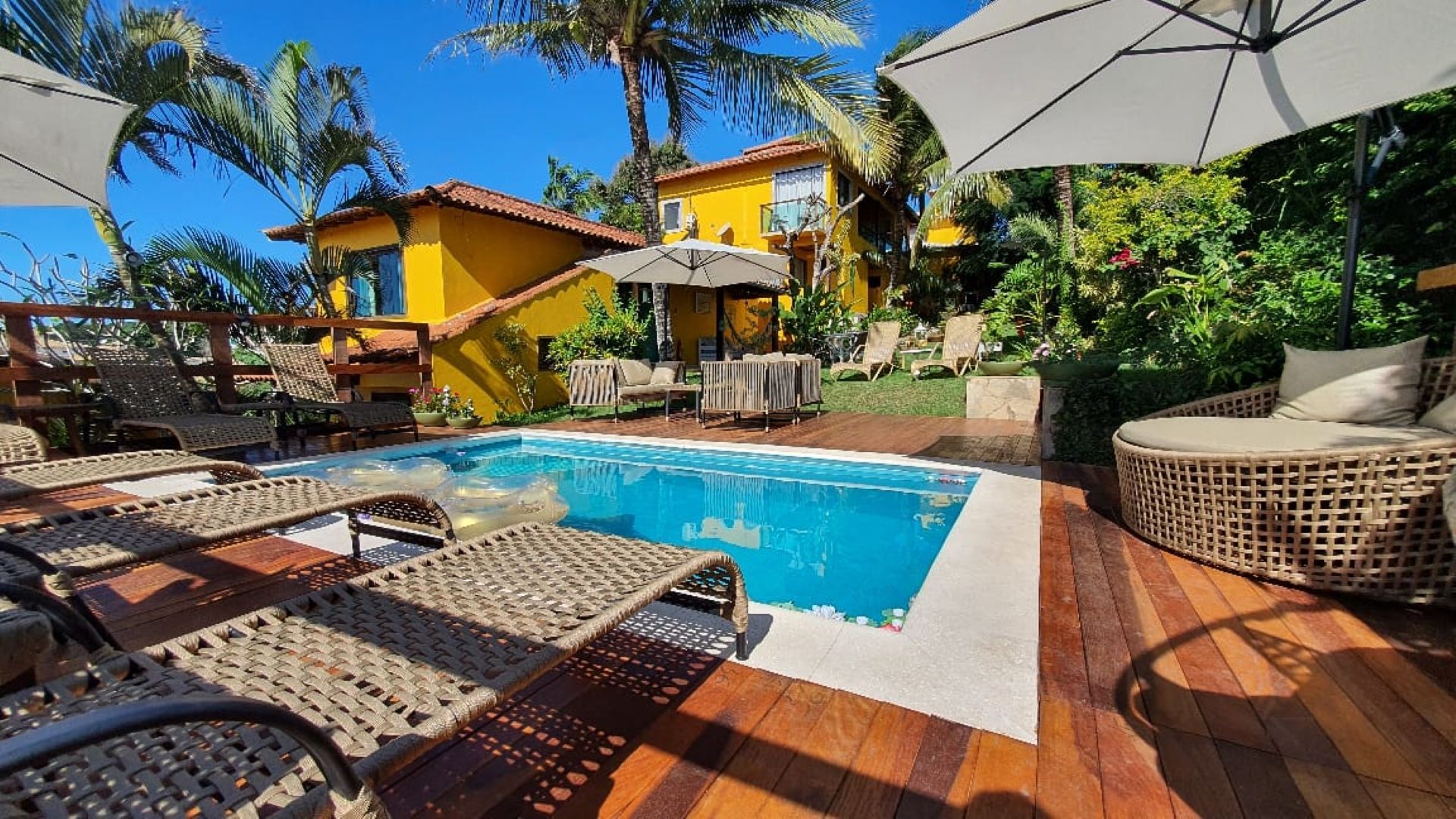 Pátio ensolarado à beira da piscina com espreguiçadeiras, guarda-sóis e uma vila amarela rodeada por plantas tropicais.